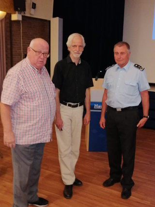 Drei Männer, einer in hellblauem Uniformhemd, stehen nebeneinander auf Holzfußboden in einem Innenraum.