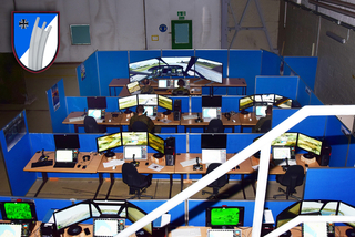 Überblicksansicht eines Militär-Simulationszentrums mit vielen Soldaten an Mehrschirm-Arbeitsplätzen für Flug-/Fahrzeugsimulationen, oben links ein Emblem.