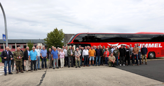 Eine große Gruppe Menschen, hauptsächlich ältere Erwachsene, steht vor einem roten Reisebus. Ein Mann in Uniform hält eine Plakette.