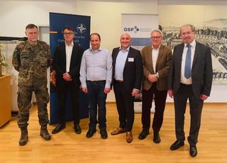 Sechs Männer, einer in Militäruniform, fünf in Zivil, stehen vor Bannern der NATO und GSP Gesellschaft für Sicherheitspolitik.