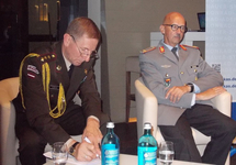 Ein lettischer Offizier unterschreibt Dokumente, während ein deutscher Offizier bei einer Veranstaltung der Konrad-Adenauer-Stiftung sitzt.