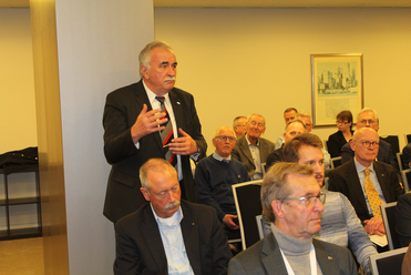Ein älterer Mann im Anzug spricht in einem Konferenzraum, während eine Gruppe ihm zuhört.