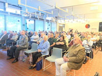 Publikum, meist ältere Erwachsene, darunter ein Mann in Uniform, sitzend in hellem Konferenzraum mit Fliesenboden.
