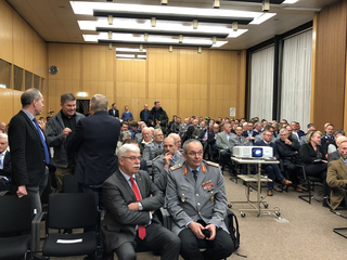 Eine große Gruppe Männer, teils in Militäruniform, bei einer Konferenz oder Versammlung in einem holzvertäfelten Raum.