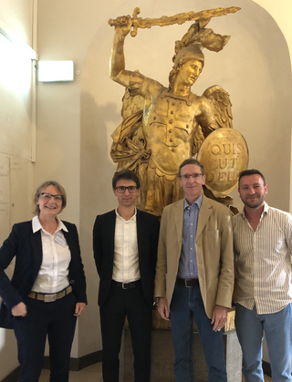 Vier Personen posieren vor einer großen goldenen Statue eines Engels, der ein Schwert und einen Schild mit der Inschrift "QUIS UT DEUS" hält.