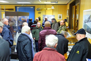 Eine Gruppe Erwachsener in einem Museum, umgeben von Vitrinen mit Artefakten und einem hängenden Modellflugzeug.