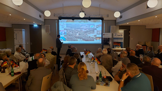 Ein Mann hält einen Vortrag vor Publikum in einem Saal, mit großer Leinwand, die eine Luftaufnahme zeigt, und deutschen Flaggen auf Tischen.