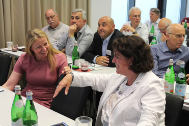 Eine vielfältige Gruppe von Fachleuten bei einem Treffen, mit zwei lächelnden und interagierenden Frauen im Vordergrund.
