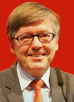 Lächelnder Mann mit Brille und braunem Haar in Anzug und oranger Krawatte vor rotem Hintergrund mit Mikrofon.
