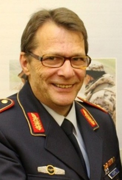 Lächelnder Mann in deutscher Militäruniform mit Brille und Brustabzeichen, vor verschwommenem militärischem Hintergrund.