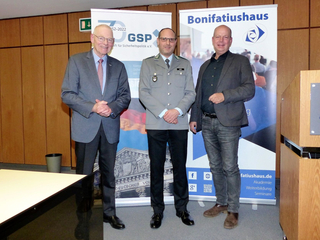 Drei Männer, darunter ein Militäroffizier, posieren lächelnd vor Bannern der GSP und des Bonifatiushaus.