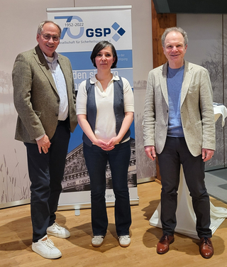 Zwei Männer und eine Frau posieren lächelnd vor einem GSP-Banner zum 70-jährigen Jubiläum (1952-2022).