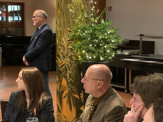 Ein Mann im Anzug steht an einer goldenen Säule und einem beleuchteten Weihnachtsbaum, während Personen im Vordergrund sitzen.