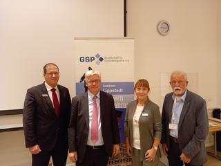 Vier Personen, drei Männer und eine Frau, posieren vor einem Banner der GSP Sektion Lippstadt bei einer Veranstaltung.