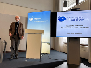 Harald Gann, Senior Advisor, präsentiert über Friedenserhaltende Maßnahmen der UN bei einem UN Bonn Event, UN-Logos auf Bildschirmen.