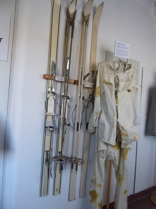 Historische Skiausrüstung mit mehreren Paar Ski, Skistöcken und einer militärischen Schneesturmkleidung an einer Wand.