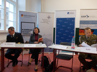 Drei Personen, darunter zwei in Uniform, sitzen an einem Tisch bei einer Podiumsdiskussion vor Veranstaltungsbannern.