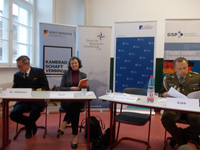 Drei Personen, darunter zwei in Uniform, sitzen an einem Tisch bei einer Podiumsdiskussion vor Veranstaltungsbannern.