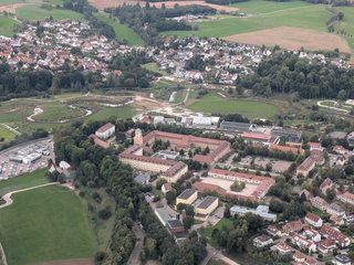 Luftaufnahme einer deutschen Stadt: Großer historischer Gebäudekomplex, Wohnsiedlungen, grüne Felder und mäandernder Fluss.
