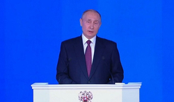 Wladimir Putin spricht an einem Rednerpult mit Mikrofonen, trägt einen dunklen Anzug und eine lila Krawatte, vor blauem Hintergrund.