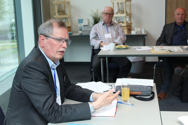 Ein Mann in Anzug und Brille gestikuliert sprechend an einem Tisch, im Hintergrund sitzen zwei weitere Männer bei einem Meeting.