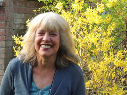 Lächelnde Frau mit blonden Haaren in blauer Strickjacke vor einem leuchtend gelben Forsythienbusch an einem sonnigen Tag.