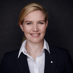 Professionelles Porträt einer lächelnden blonden Frau mit blauen Augen, die einen dunklen Anzug und ein weißes Hemd trägt.