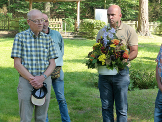 Ein kahler Mann hält ein großes, farbenfrohes Blumenarrangement im Freien, flankiert von zwei weiteren Männern auf einer Grasfläche.
