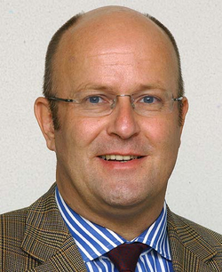 Porträt eines lächelnden Mannes mit Brille, Glatze, gestreiftem Hemd und kariertem Sakko.