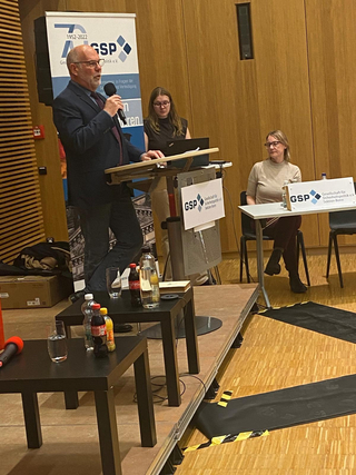 Mann spricht bei einer GSP-Sicherheitspolitik-Veranstaltung, mit zwei Frauen und GSP Bonn Schildern.