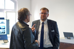 Zwei Männer unterhalten sich in einem hellen Raum. Ein Mann im Anzug hört einem Mann in Lederjacke zu, der gestikuliert.