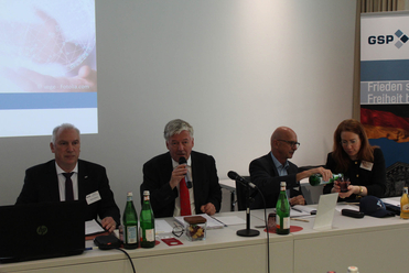 Vier Personen bei einer Konferenz; ein Mann spricht ins Mikrofon, ein anderer schenkt einer Frau ein Getränk ein.