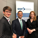 Drei lächelnde Personen, zwei Männer und eine Frau, vor einem Banner der GSP Gesellschaft für Sicherheitspolitik e.V.