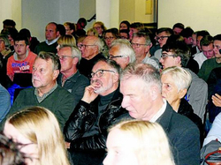 Ein diverses Publikum von Erwachsenen und einigen jüngeren Leuten sitzt aufmerksam in einem Raum und blickt nach vorne.