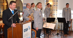 Ein Mann spricht an einem Pult mit Mikrofon, während eine Militärblaskapelle in grauen Uniformen hinter ihm steht.
