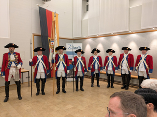 Männer in historischen rot-blauen Uniformen mit Dreispitzen, teils mit Fahnen oder Speeren, stehen bei einer Veranstaltung.