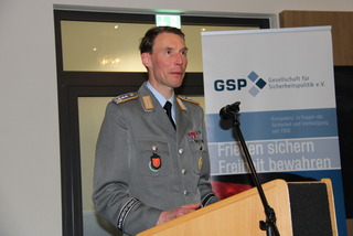 Dr. Freuding, ein Mann in Militäruniform, spricht an einem Rednerpult vor einem Banner der Gesellschaft für Sicherheitspolitik.