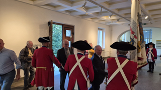 Männer in roten historischen Uniformen und Dreispitzenhüten stehen mit Zivilisten und einer großen Zeremonialflagge drinnen.