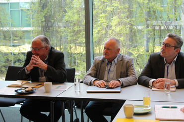 Drei Männer in Geschäftskleidung sitzen an einem Tisch in einem Meeting, mit Notizbüchern, Getränken und Blick ins Grüne.