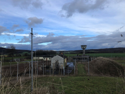 Ein verlassener Militärkomplex mit Wachturm, Gebäuden und Zäunen in einer ländlichen Landschaft unter bewölktem Himmel.