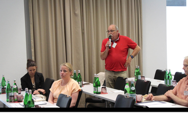Mann spricht ins Mikrofon bei einer Konferenz, Teilnehmer sitzen an Tischen mit Wasserflaschen.