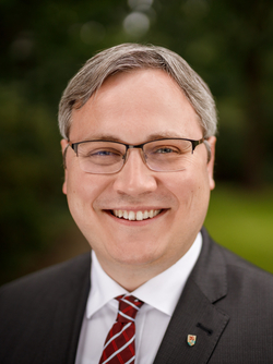 Ein lächelnder Mann mit grauem Haar und Brille in Anzug und gestreifter Krawatte blickt in die Kamera, grüner Hintergrund.