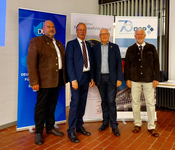 Vier Männer, teils in Anzügen, teils in Trachtenjacken, lächeln vor Roll-up-Bannern deutscher Organisationen.