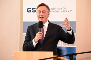 Ein Mann im Anzug spricht lächelnd in ein Mikrofon, gestikulierend vor einem Banner der "Gesellschaft für Sicherheitspolitik e.V.".