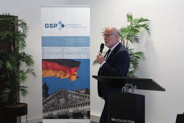 Mann spricht mit Mikrofon vor einem GSP-Banner mit deutscher Flagge, Reichstagsgebäude und Text 'Frieden sichern Freiheit bewahren'.