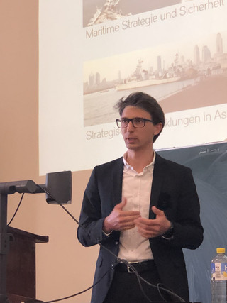 Ein Mann mit Brille hält einen Vortrag über maritime Strategie und Sicherheit vor einer Projektion mit Stadtansicht und Schiff.