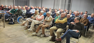 Eine diverse Gruppe meist älterer Erwachsener sitzt aufmerksam in Reihen von Stühlen bei einer Indoor-Veranstaltung. Einige nutzen Rollstühle oder Gehstöcke.