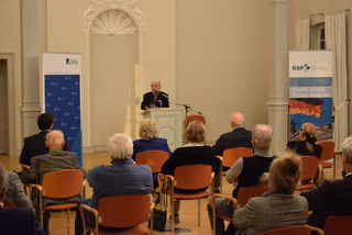 Ein Mann spricht an einem Pult vor einem Publikum, mit Bannern der Konrad-Adenauer-Stiftung und GSP im Hintergrund.