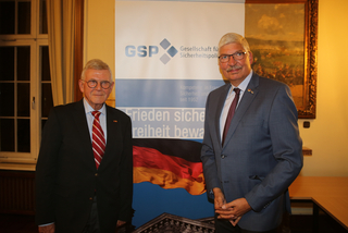 Zwei ältere Männer in Anzügen stehen vor einem GSP-Banner mit der Deutschlandflagge und dem Reichstagsgebäude.