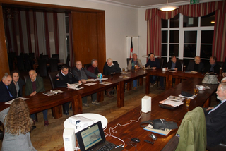 Eine Gruppe von Menschen bei einem Treffen an U-förmig angeordneten Holztischen in einem Raum, mit einem Laptop und Beamer.